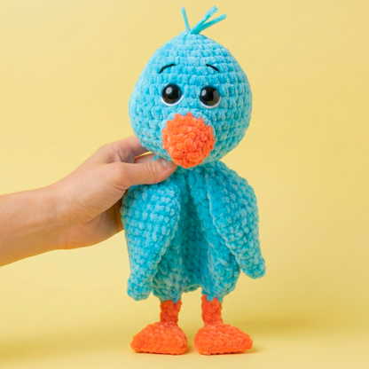 Hand holding a blue and orange crocheted plush bird toy with large eyes on yellow background
