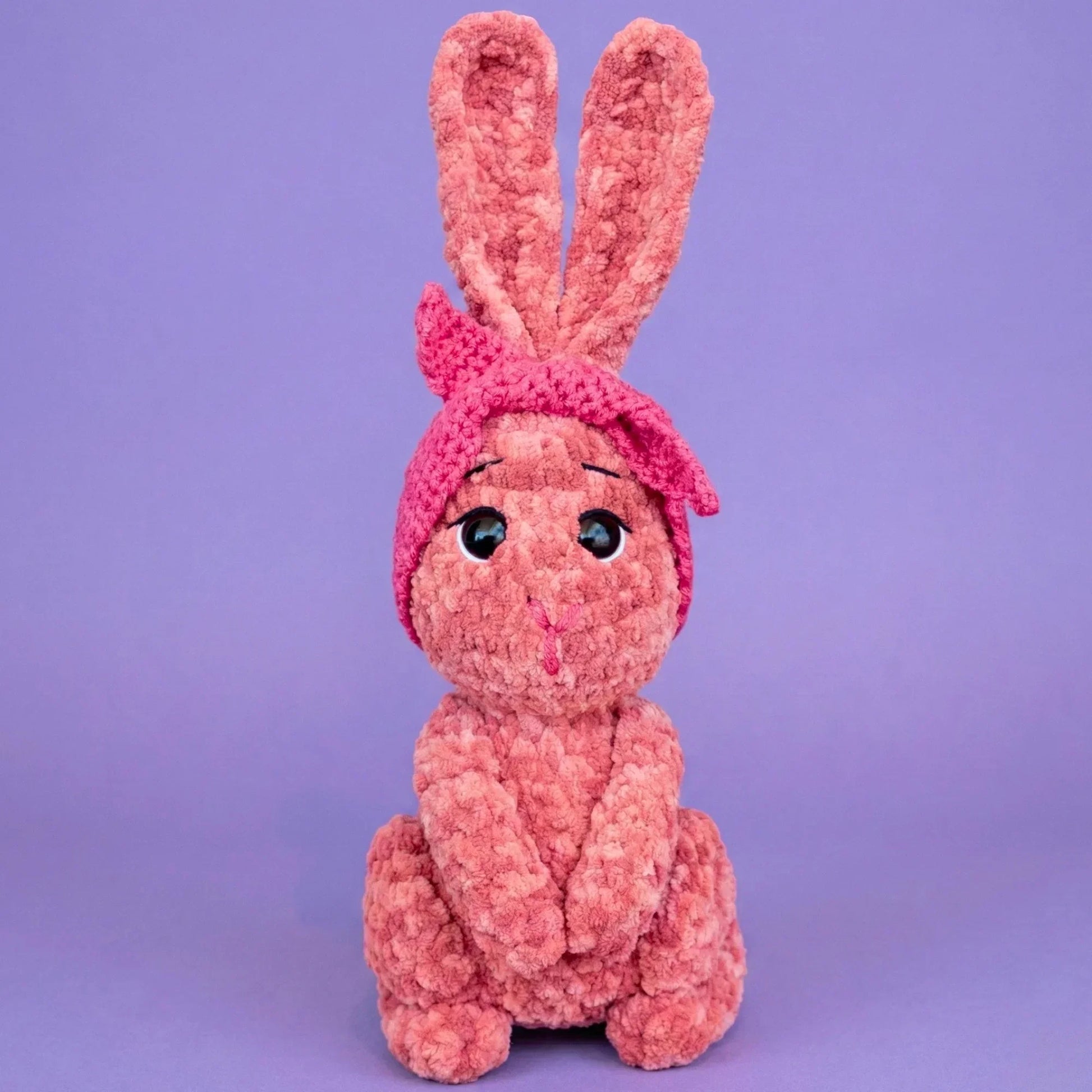 Pink crochet bunny plushie with big eyes and headband, amigurumi pattern on purple background