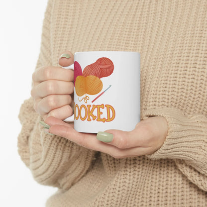 Person holding a mug with yarn and knitting needles design and 'HOOKED' text.