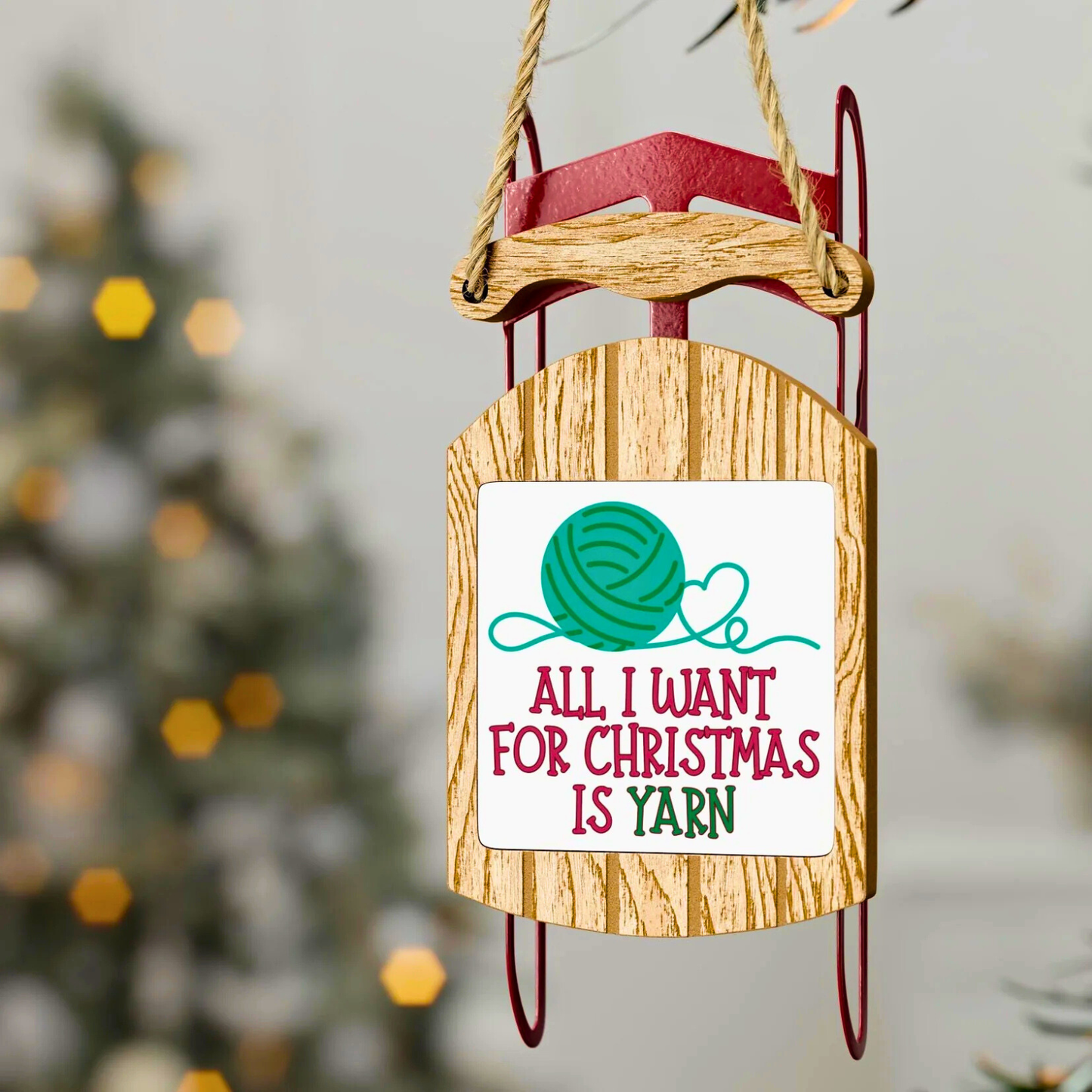Wooden sled ornament with a Christmas message and yarn graphic, blurred Christmas tree in the background.