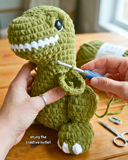 Green crochet dinosaur being worked on with a crochet hook on a wooden surface.