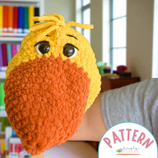 Crocheted orange and yellow bird head with 'Simply Hooked' pattern label in a room setting.
