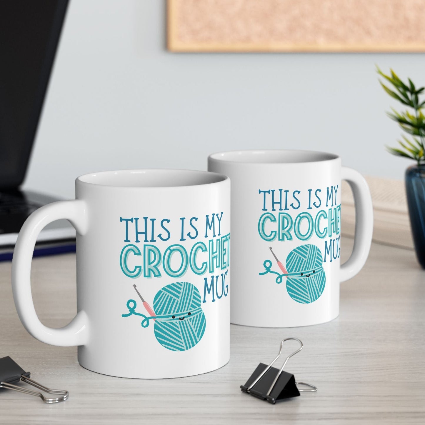 Two white mugs with teal text and yarn ball graphics on a desk with office supplies.
