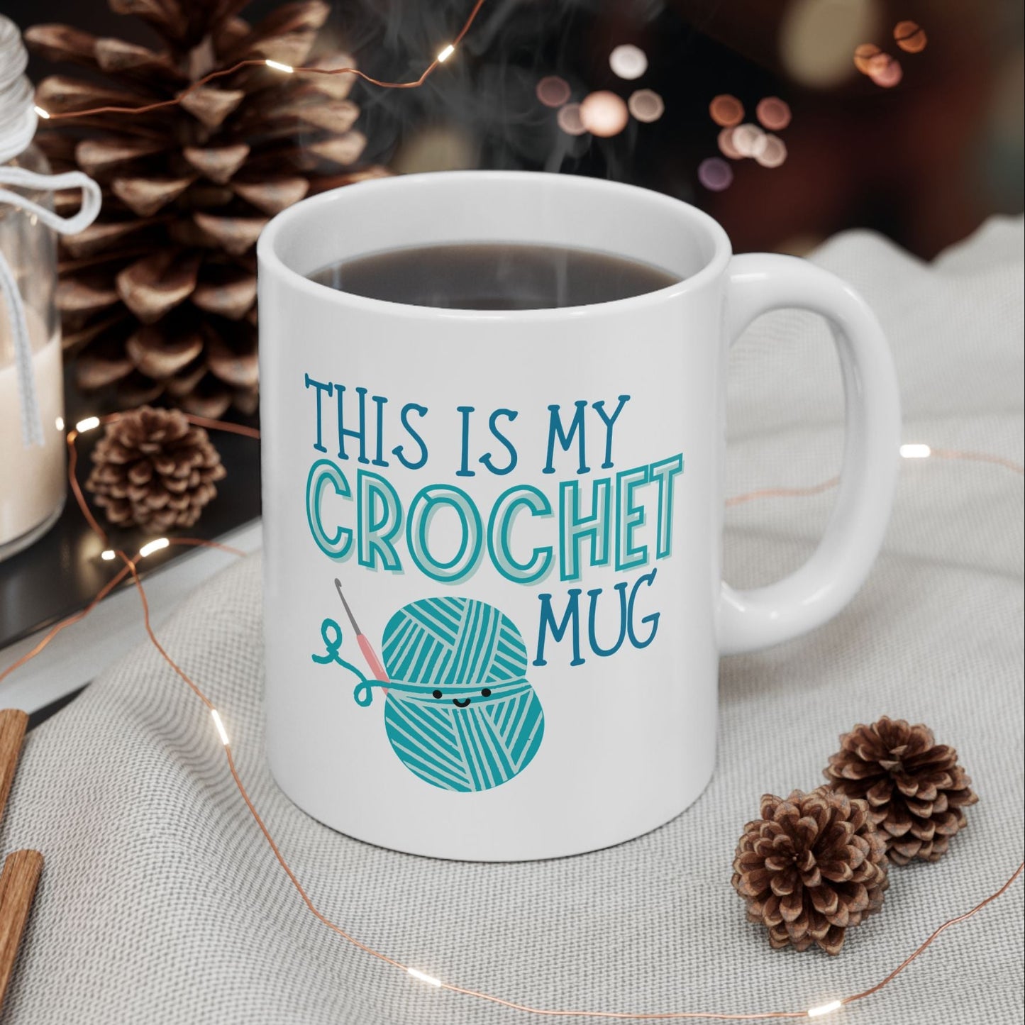 White mug with 'This is my crochet mug' text and yarn ball graphic, surrounded by decorative items on a table.