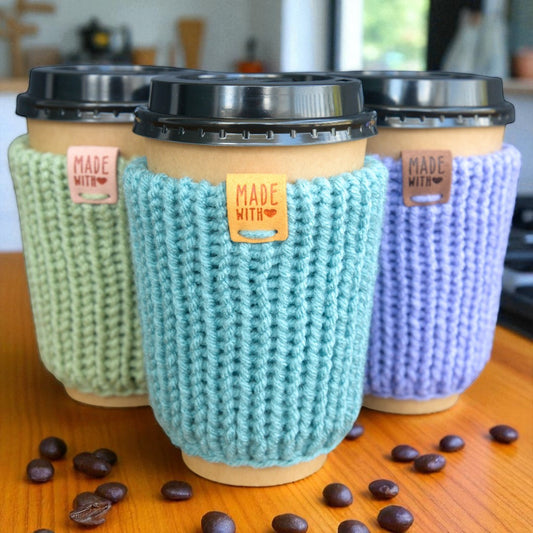 Three coffee cups with knitted cozies in green, blue, and purple on a wooden surface with coffee beans.