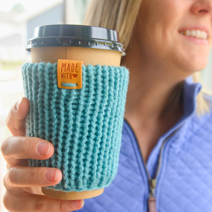 Person holding a coffee cup with a teal knitted sleeve and 'Made with Love' tag.