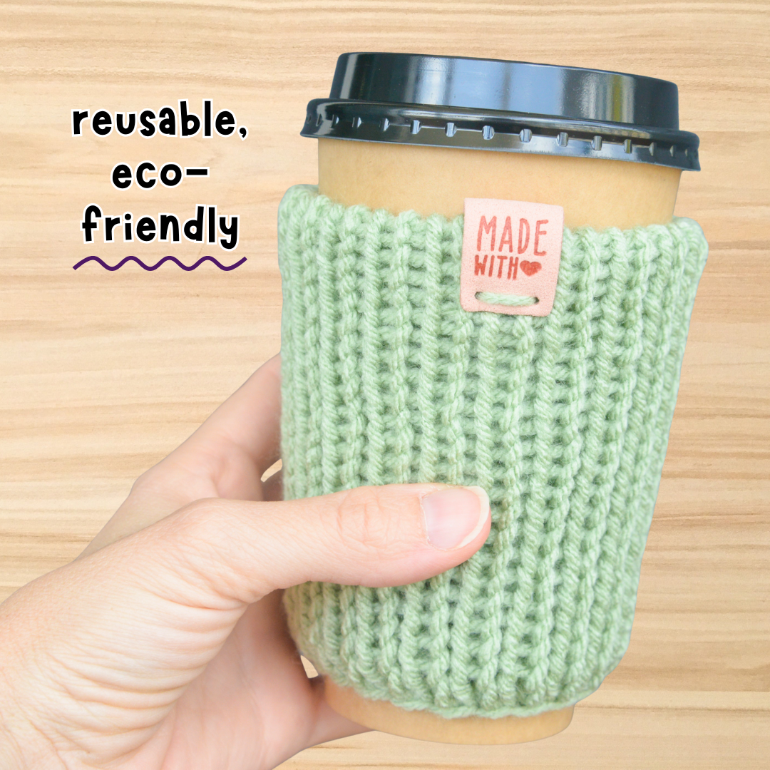 Green knitted coffee cup sleeve held by a hand with text 'reusable, eco-friendly' on a wooden background.