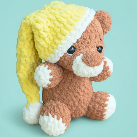 Plush brown teddy bear with white accents wearing a yellow knitted nightcap against light blue background