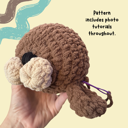A hand holds a crocheted plush toy with a brown body, cream-colored snout and feet, and a black eye, against a pale yellow background with decorative brushstroke graphics and text stating 'Pattern includes photo tutorials throughout.'.