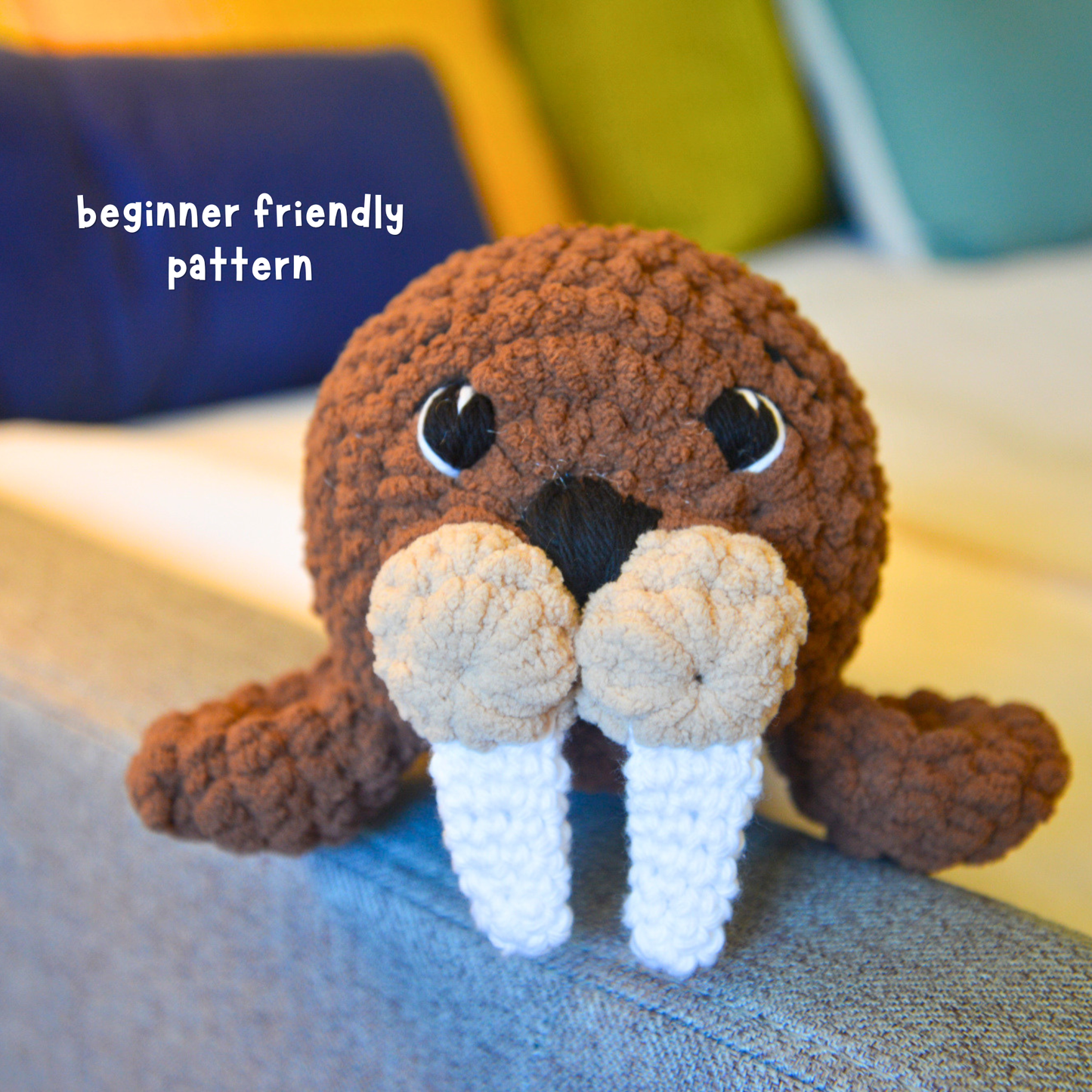 A crocheted plush walrus toy sitting on a blue-gray couch with colorful pillows in the background. White text in the upper left reads 'beginner friendly pattern'.