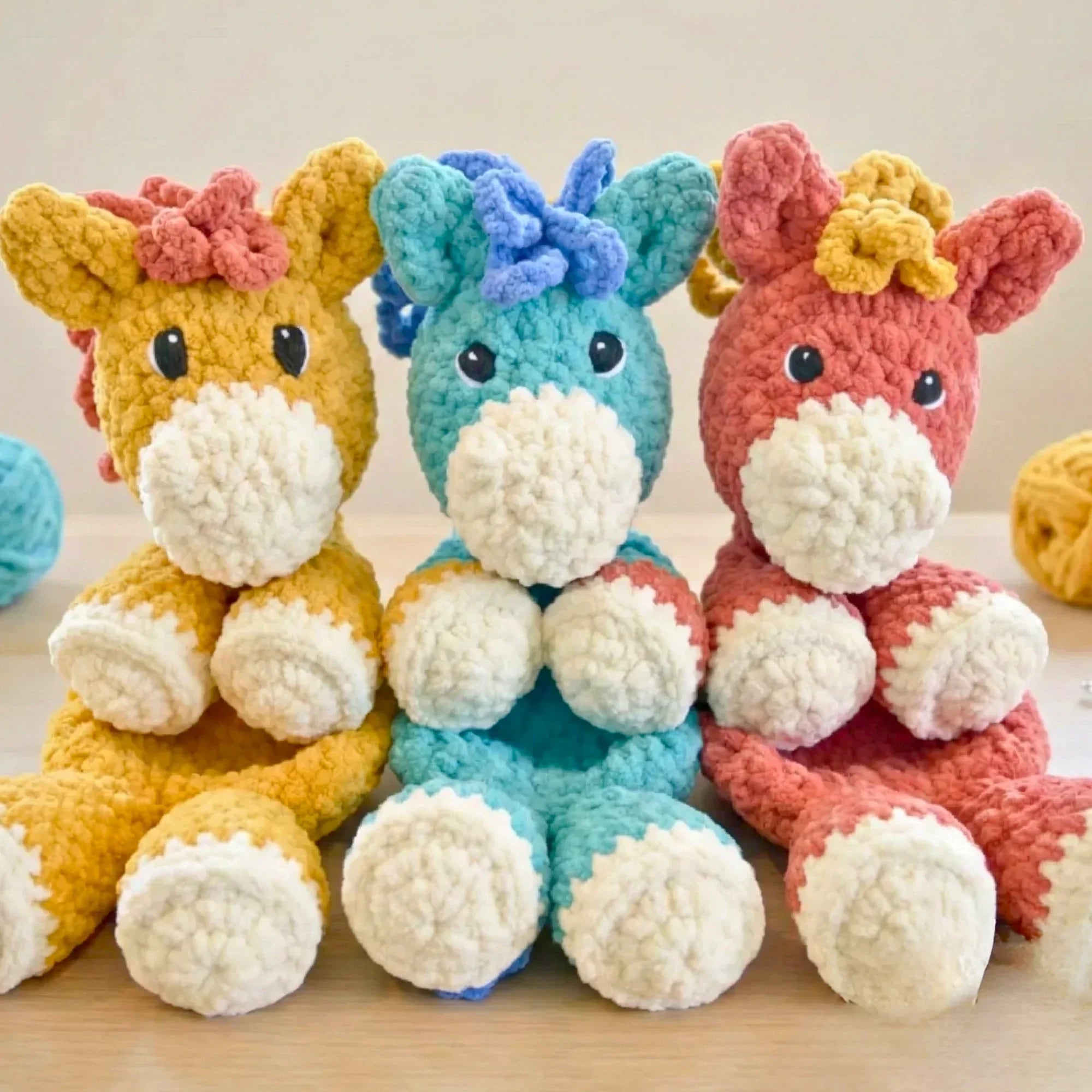 Three plush crochet horses in yellow, blue, and coral, displayed on a wooden table.