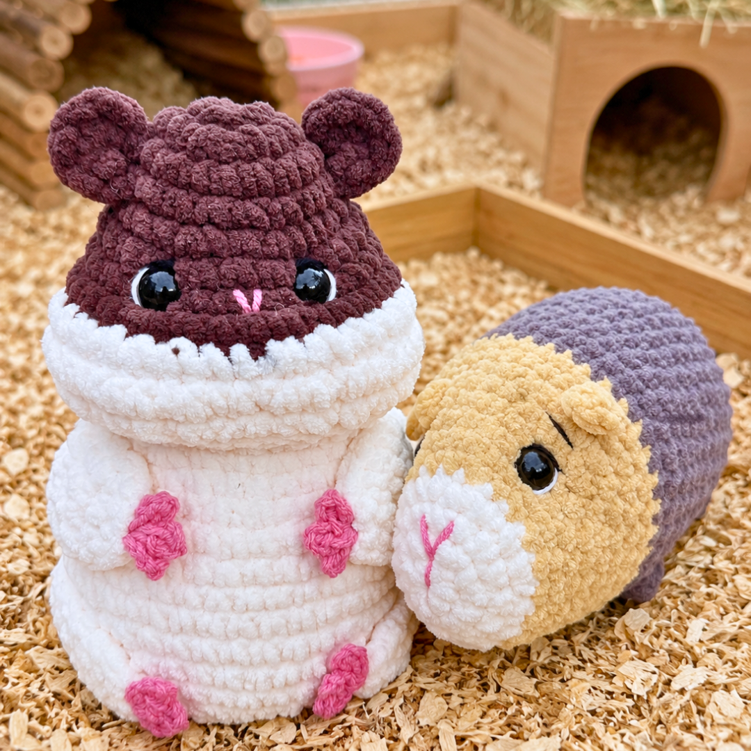 Two crocheted hamster toys placed on wood shavings in a small hamster enclosure. One toy is shaped like a white hamster wearing a dark plum hood, and the other is a round hamster figure with a tan face, white belly, and purple hood.