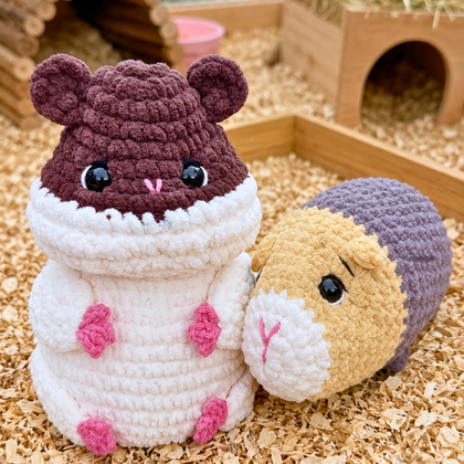 Two crocheted hamster toys placed on wood shavings in a small hamster enclosure. One toy is shaped like a white hamster wearing a dark plum hood, and the other is a round hamster figure with a tan face, white belly, and purple hood.