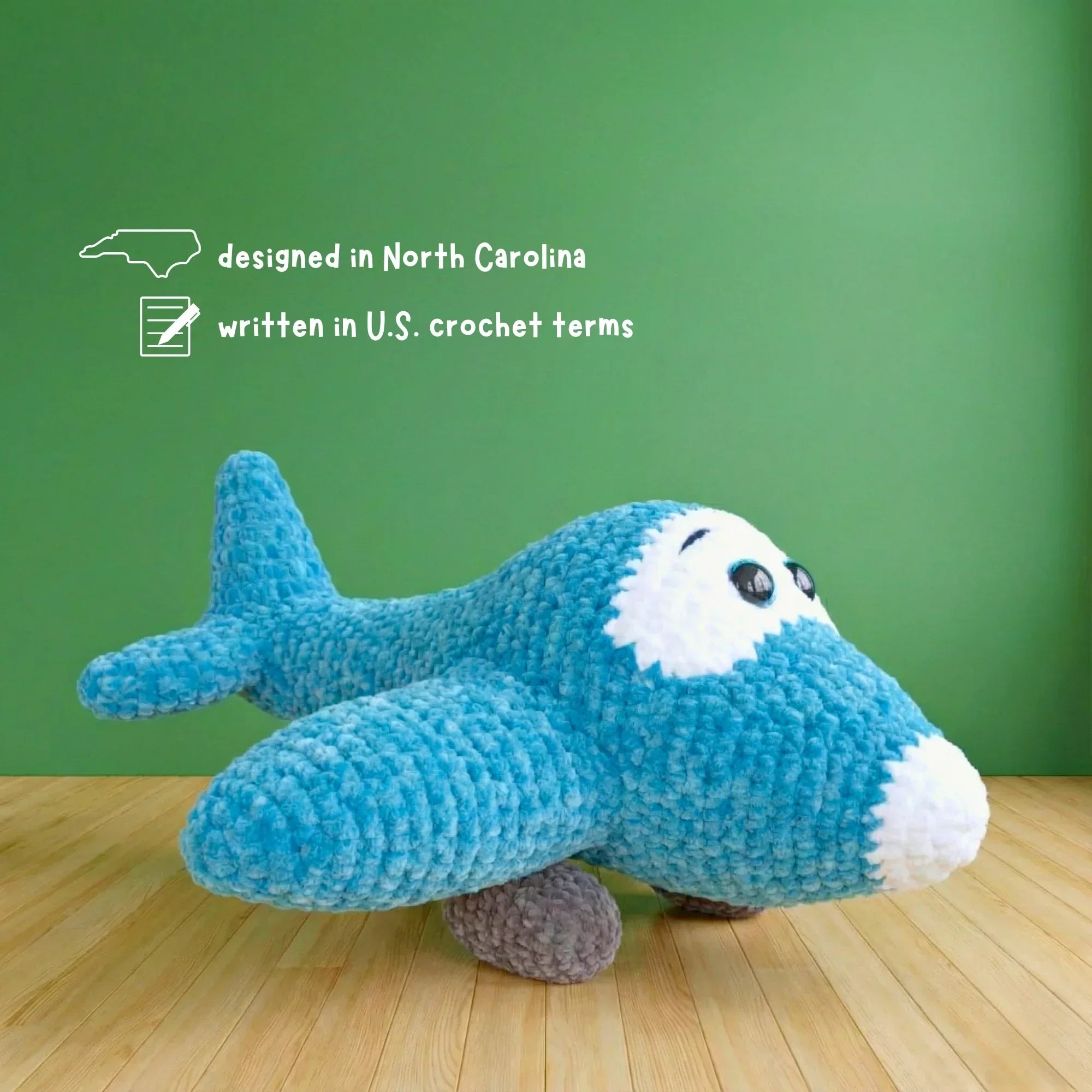 Blue crochet airplane plush amigurumi with white face and black eyes on wood floor, green background.