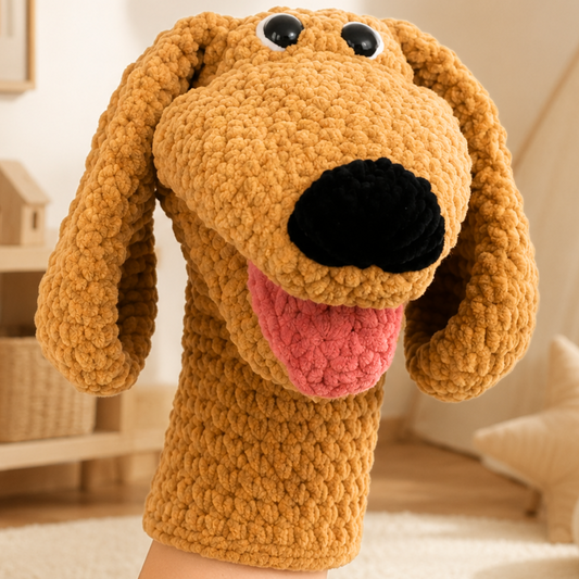 A close-up of a crocheted dog-shaped hand puppet with long floppy ears, black eyes, a black nose, and a pink tongue, shown against a softly blurred indoor room background.