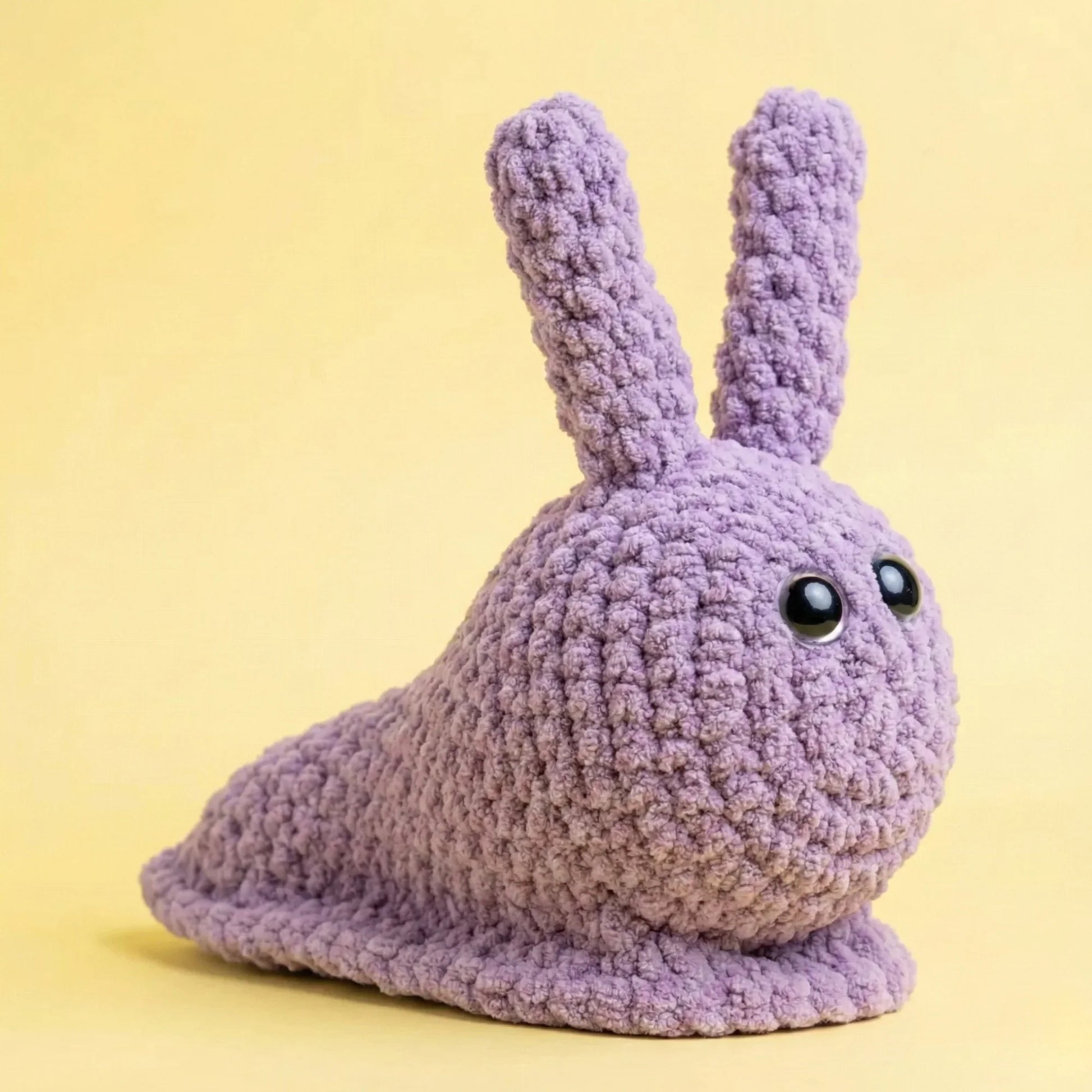 Purple crochet snail plushie with black eyes on a yellow background, amigurumi pattern