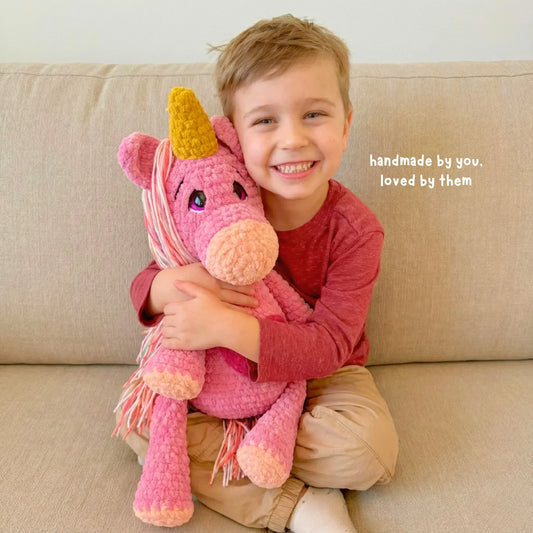 Smiling child hugging a pink crochet unicorn plushie with a yellow horn on a beige sofa